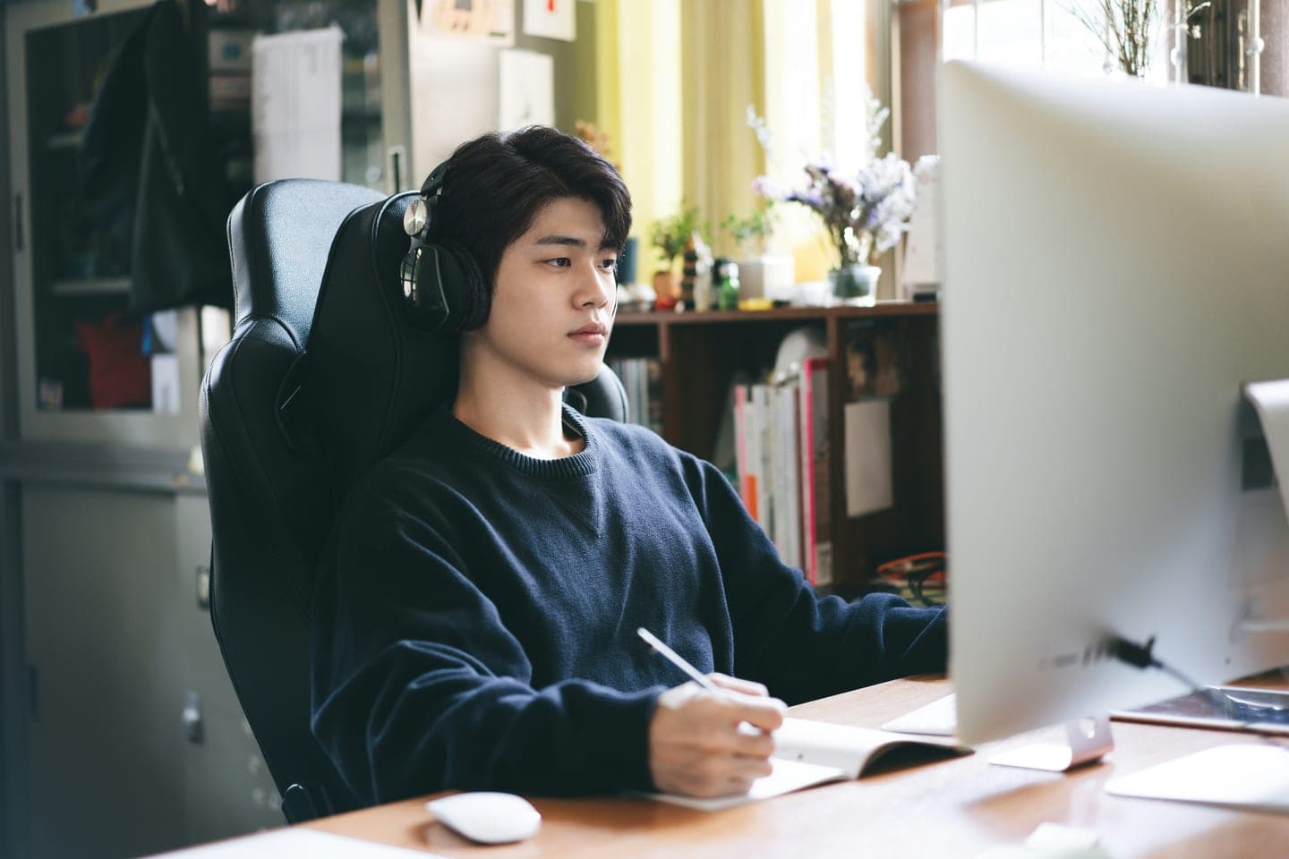 헤드폰을 쓰고 큰 모니터 화면을 보며 공책에 받아 적는 온라인 수업 장면